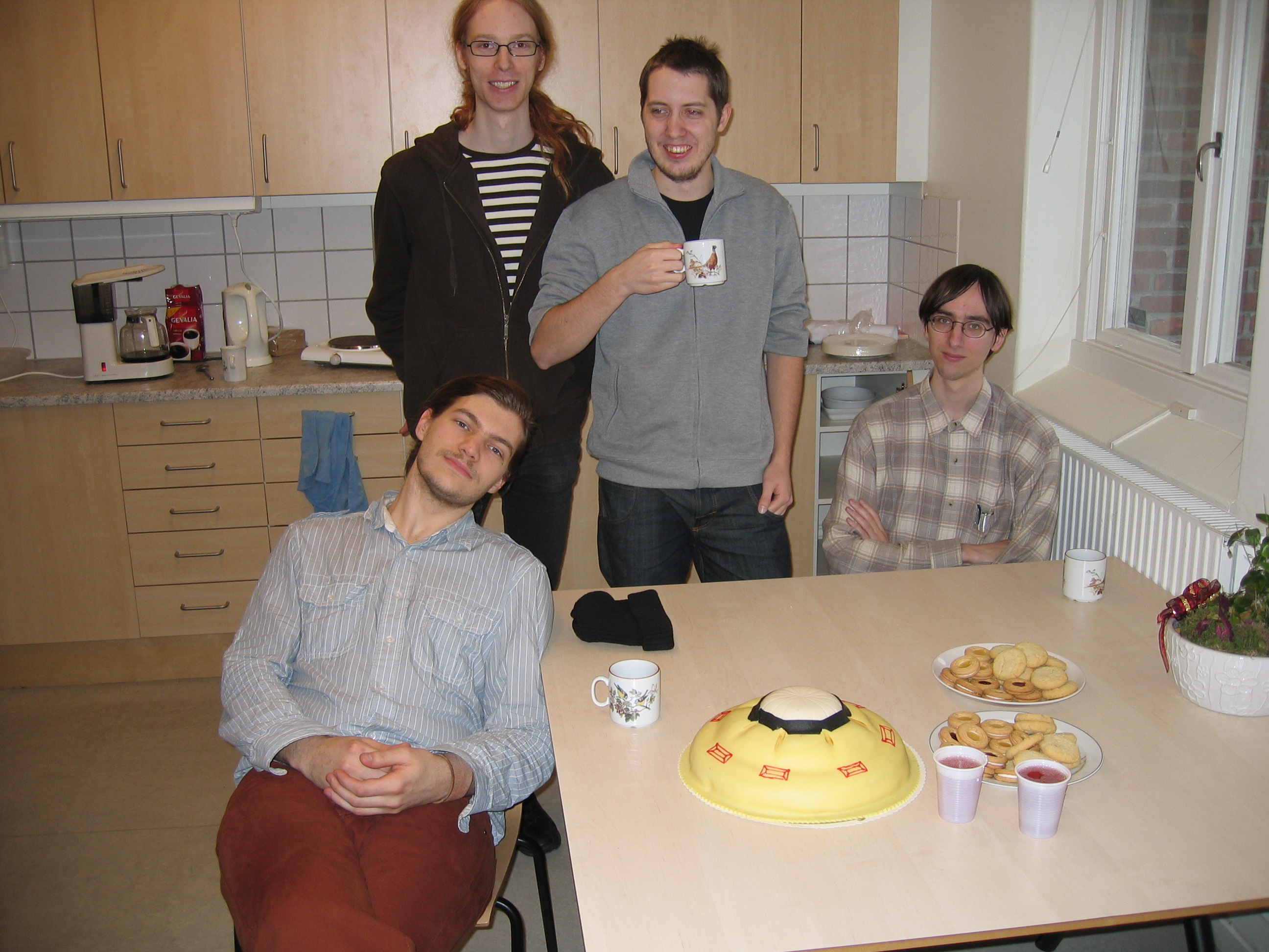 Alexander, Daniel, Jens, and Jonas all facing the camera and seated around a table with a cake shaped like the D-70 enemy from Harvest: Massive Encounter.